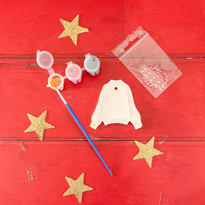 Illustration d’un kit créatif contenant une suspension en argile en forme de pull de Noël, avec peintures, vernis et strass.