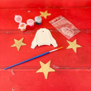 Illustration d’un kit créatif contenant une suspension en argile en forme de pull de Noël, avec peintures, vernis et strass.